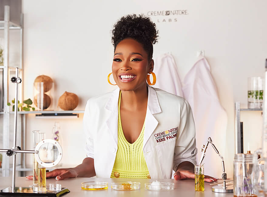 Keke Palmer, Chief Brand Officer for Creme of Nature in a lab coat sitting at a chemist's table